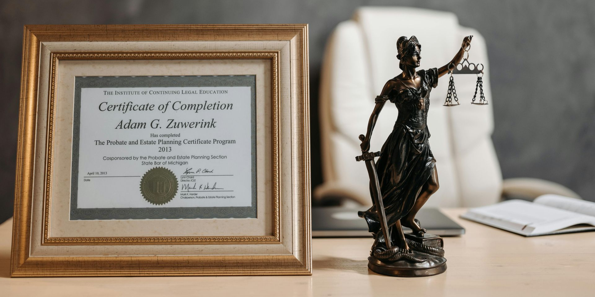 Framed certificate beside a bronze Lady Justice statue on a desk with a laptop in the background