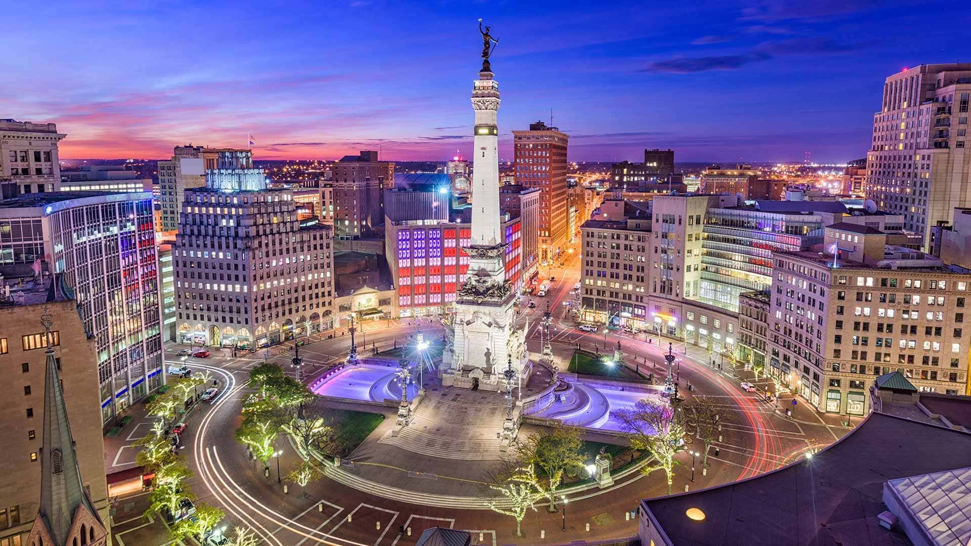 City square at dusk with a lit monument, colorful fountains, and surrounding buildings in the skyline