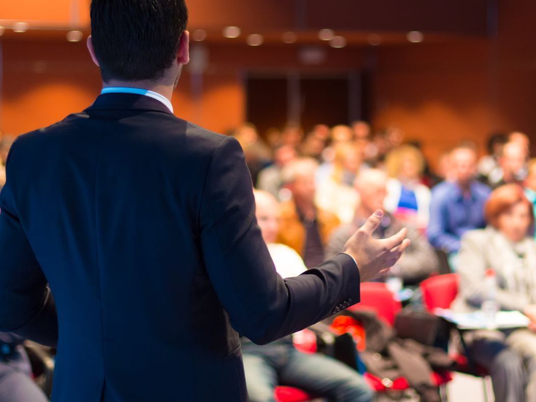 Speaker in dark suit addressing a seated audience in a conference hall