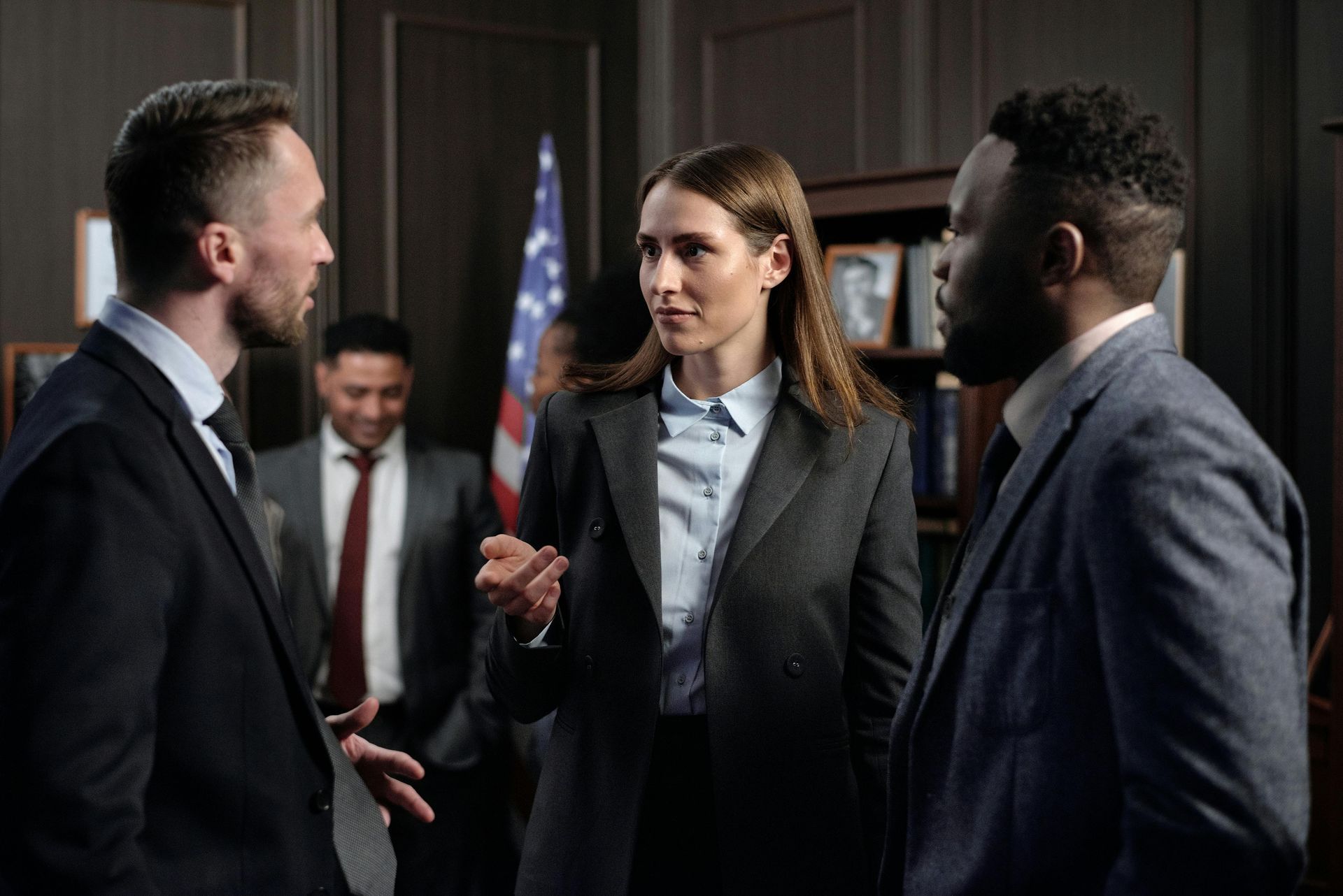 Three professionals in suits talking in a dim conference room, with one man blurred in the background.