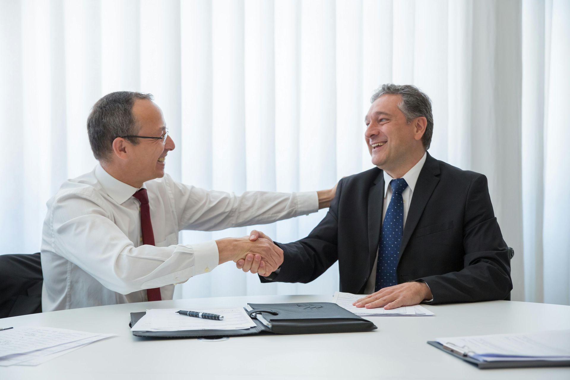 Two businessmen shaking hands across a white conference table in a bright office