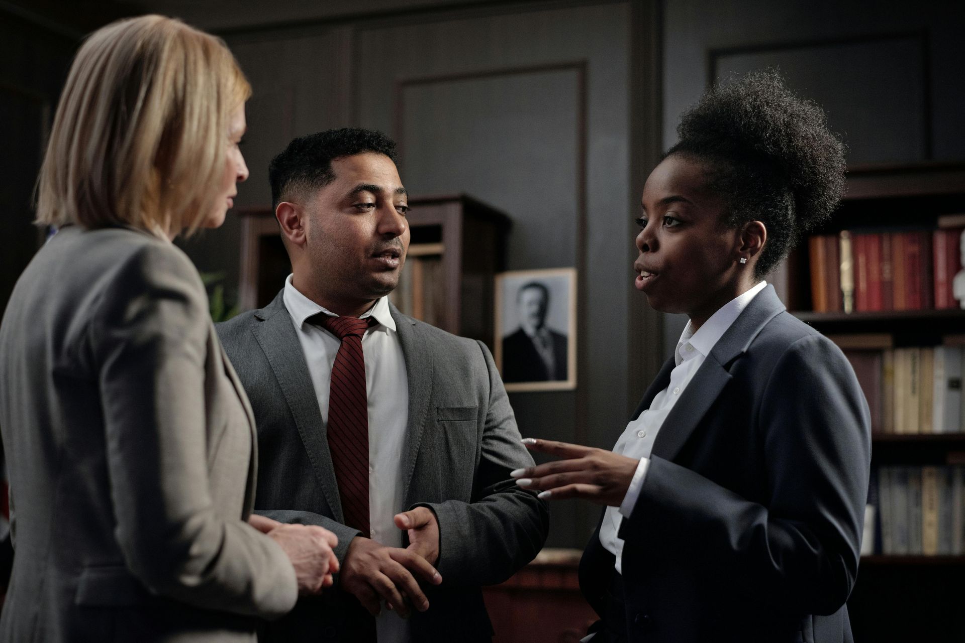 Three coworkers in business attire talking in an office, one holding a tablet.
