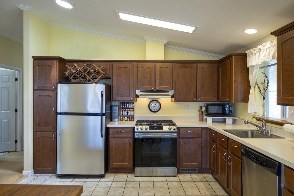 A large, tiled eat-in kitchen in a house. Brown stained wooden cabinets, stainless-steel refrigerator, gas range, microwave, dishwasher.