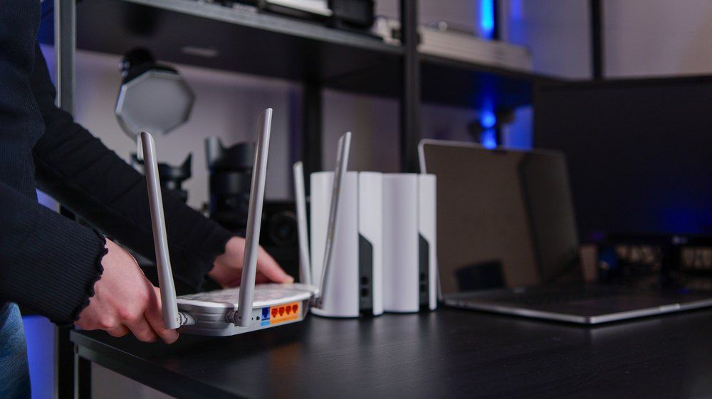Hands adjusting a wireless router with mesh network devices and laptop in background