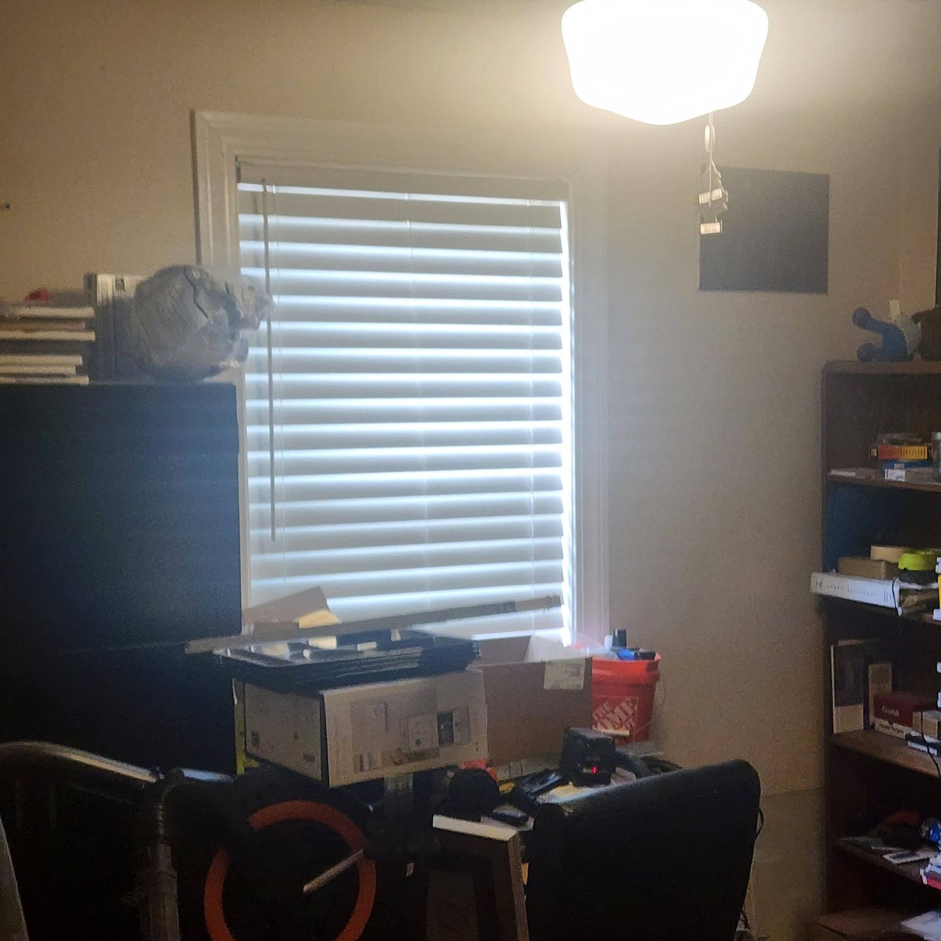 Cluttered home office with desk, bookshelves, window blinds, and ceiling light.