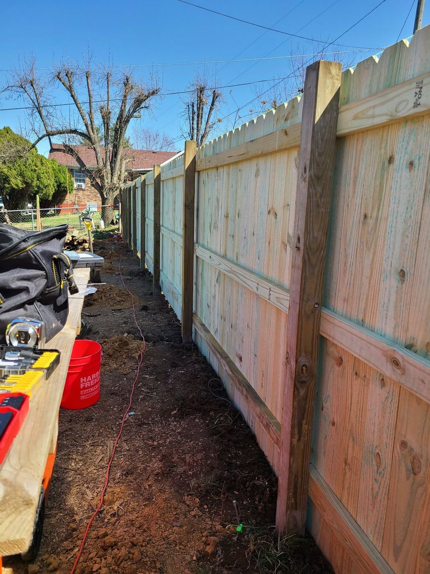 Narrow side yard with a wooden fence, dirt path, and outdoor tools under a clear blue sky.