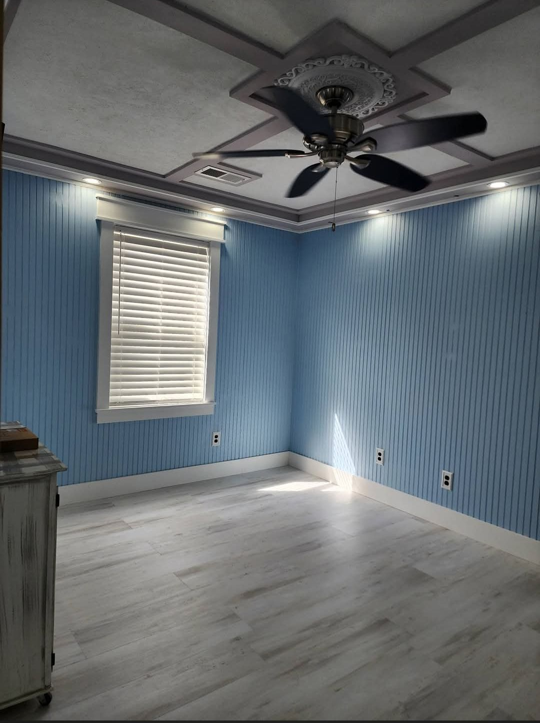 Empty blue room with white trim, light wood floor, ceiling fan, and a window with blinds.