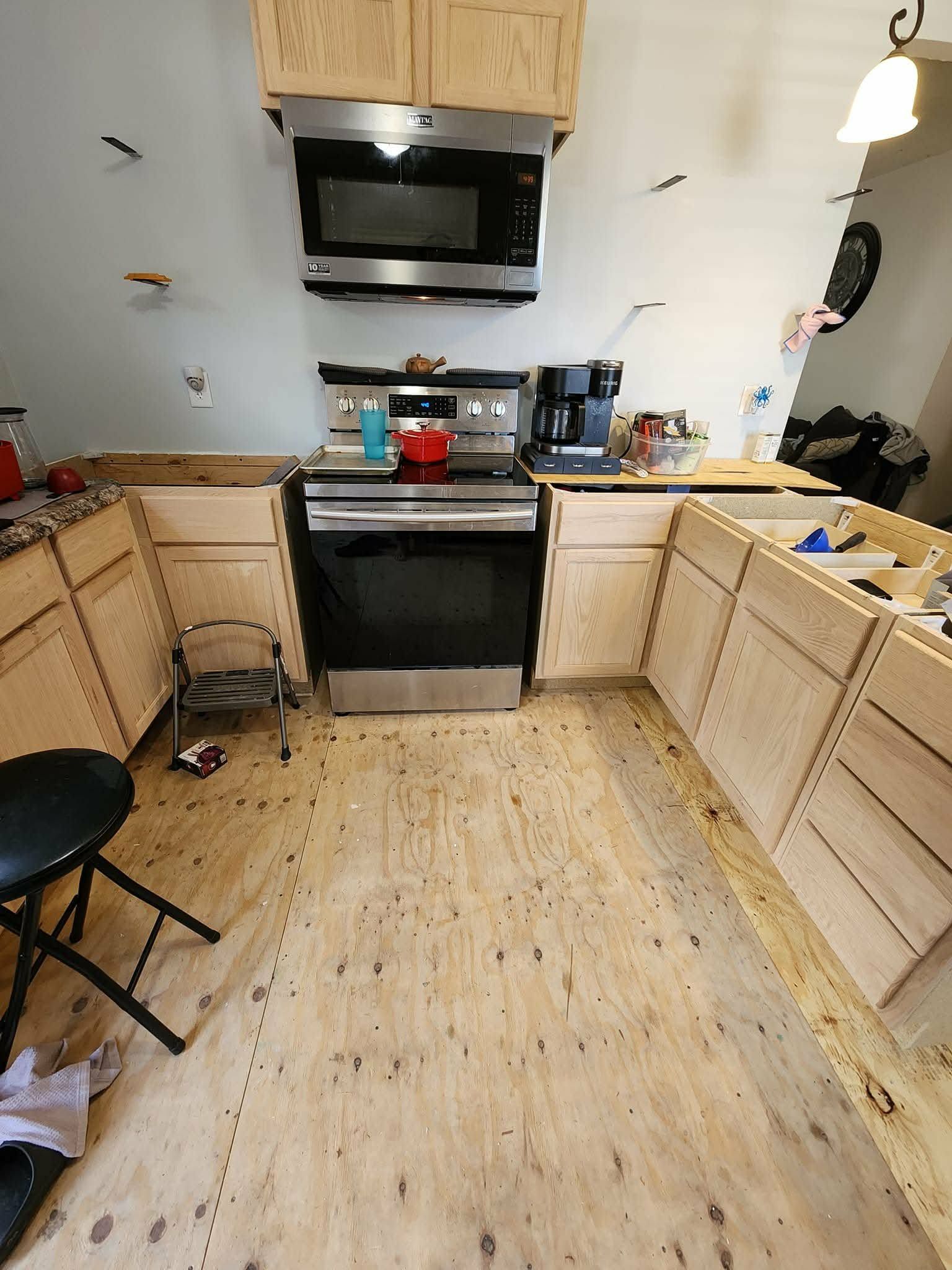 Small kitchen with light wood cabinets, black stove and microwave, and a cluttered floor and countertops.