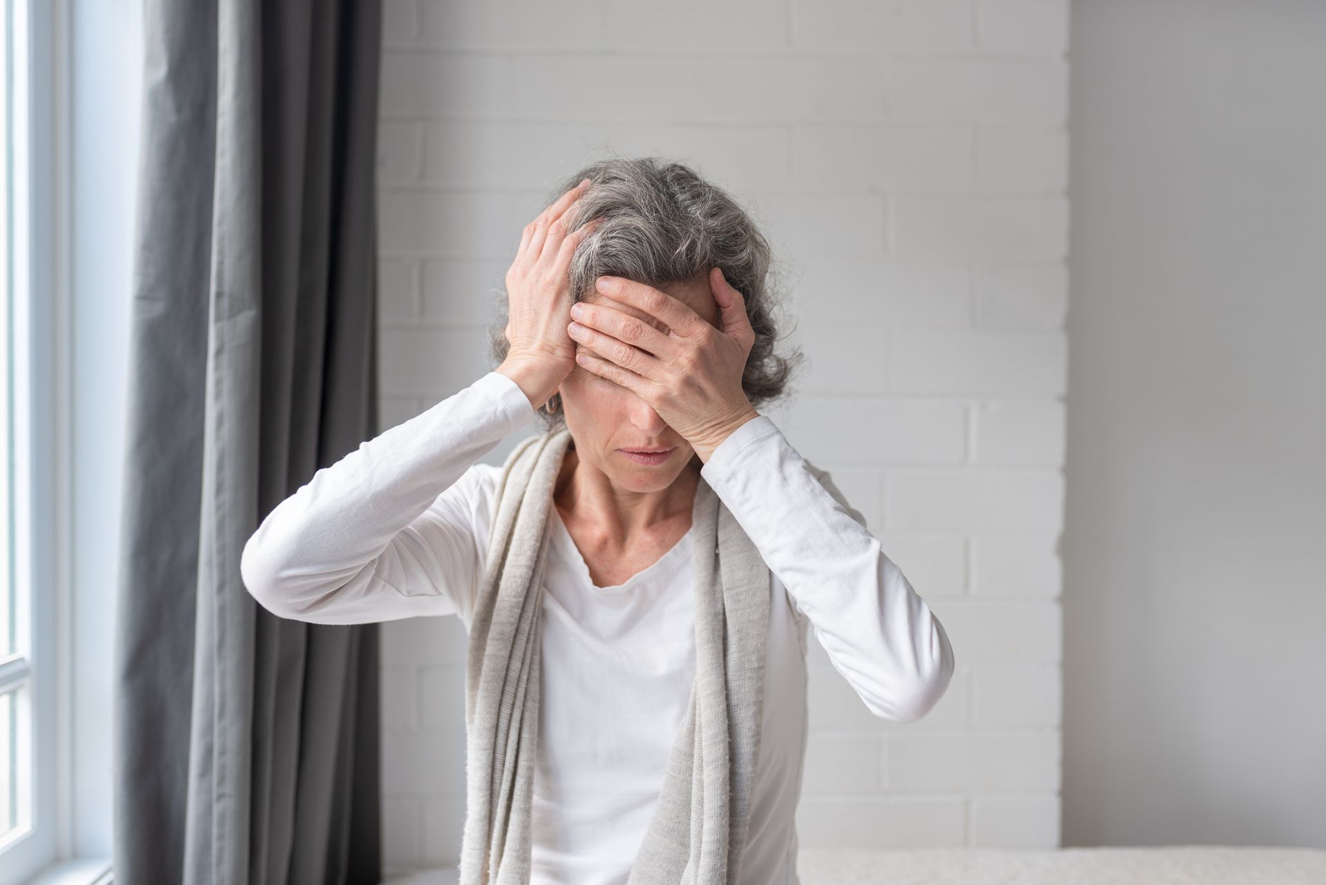 Woman with gray hair, hands on forehead, appears stressed or in pain, in a room with a window.