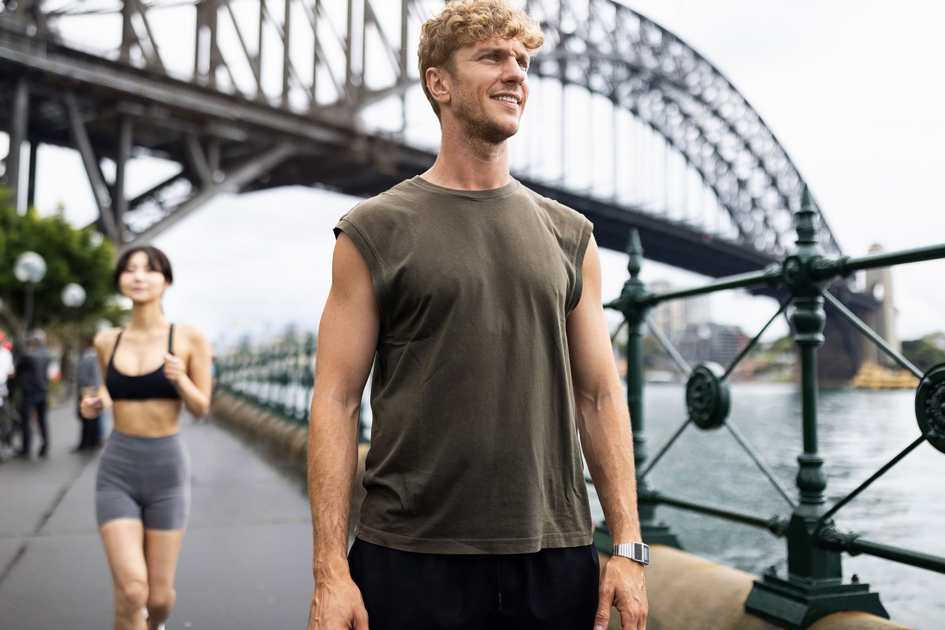Man in a sleeveless shirt smiles near Sydney Harbour Bridge; woman runs past in the background.