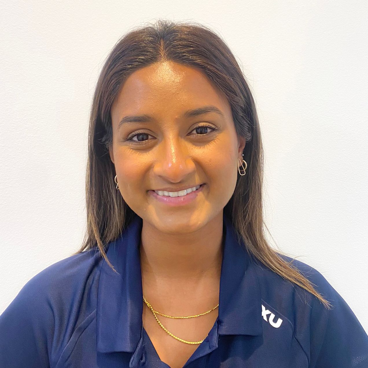 Woman smiling, wearing a dark blue shirt and gold necklace, in front of a white wall.
