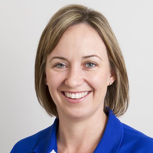 Woman with short brown hair, smiling, wearing a blue jacket, against a white background.