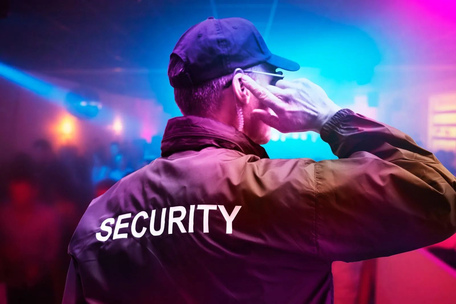 Security guard wearing a hat and jacket, illuminated in purple and blue lights, talking into a radio earpiece.