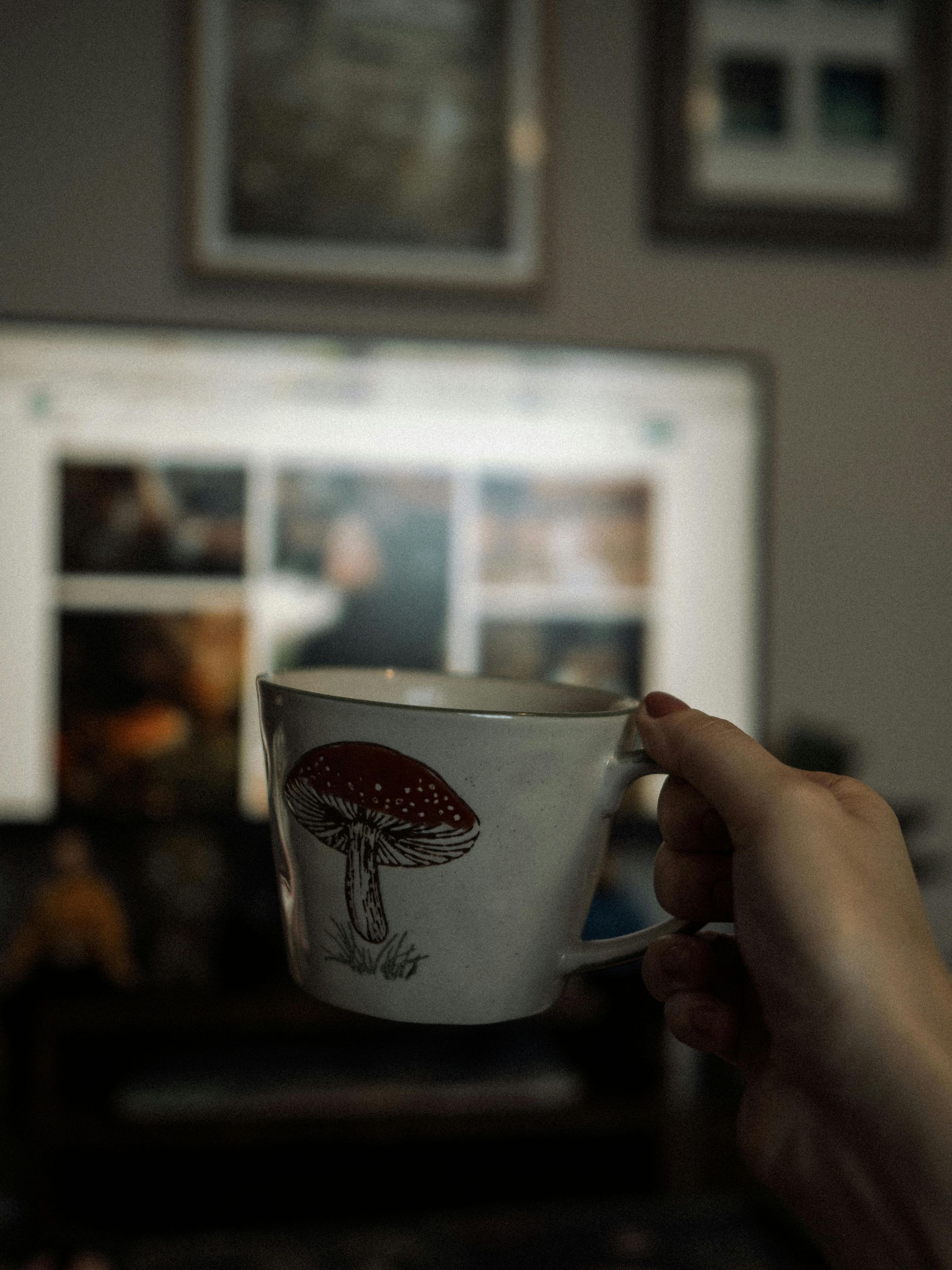A hand holds a white mug with a red mushroom illustration in front of a softly blurred computer screen.