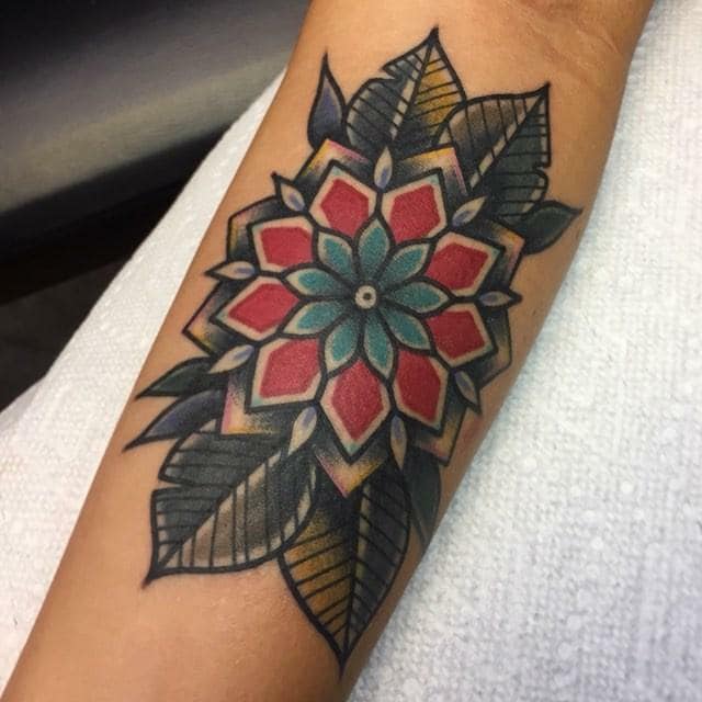 Colorful mandala-style flower tattoo with red, teal, black, and gray leaves on a forearm
