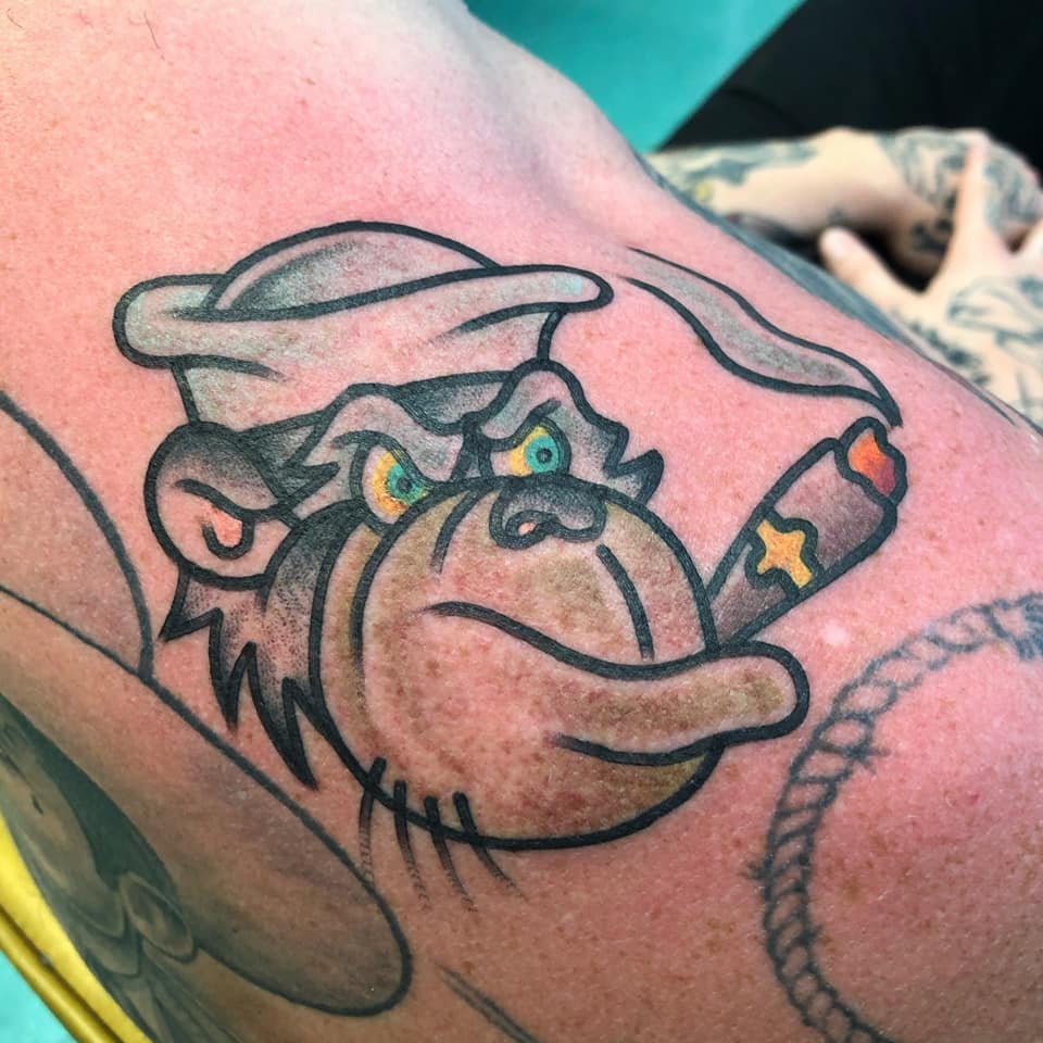Cartoon monkey tattoo with a hat and cigarette, outlined in black with yellow eyes and pink skin accents