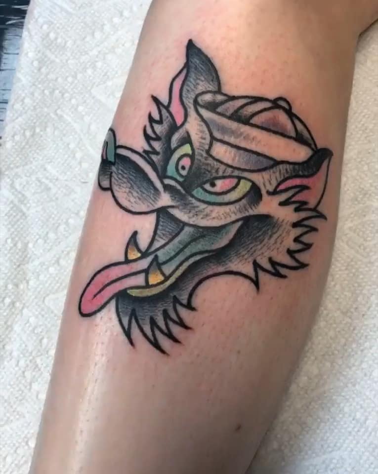 Colorful cartoon wolf tattoo with green eyes and tongue out on a person’s forearm
