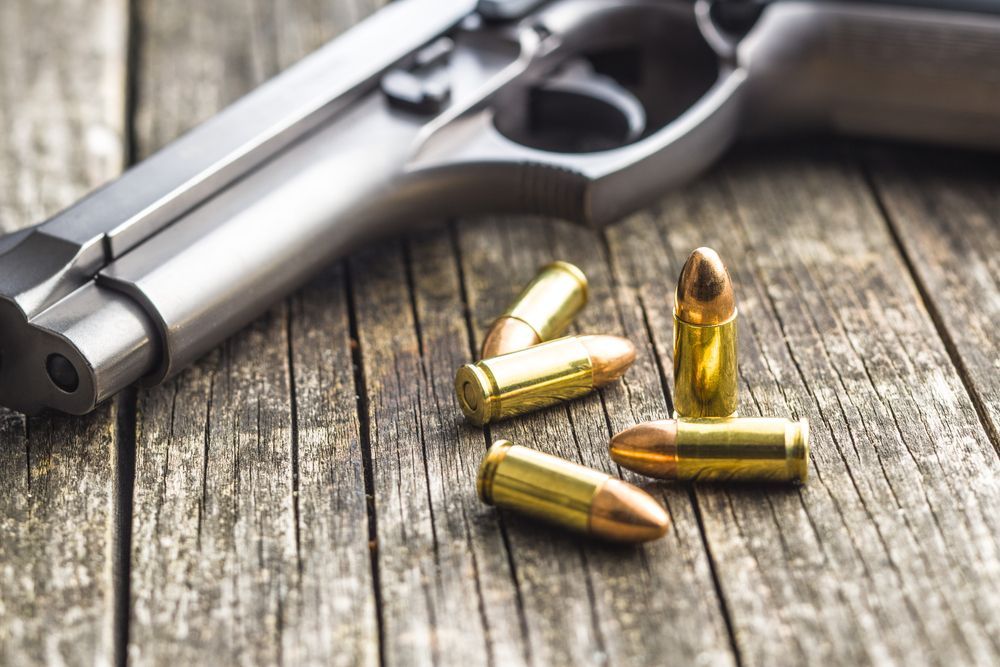 A metallic handgun rests on a textured wooden surface next to four brass-cased bullets.