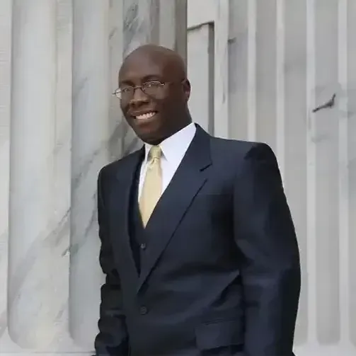 A smiling person in a dark suit, vest, and gold tie standing against a background of white marble columns.