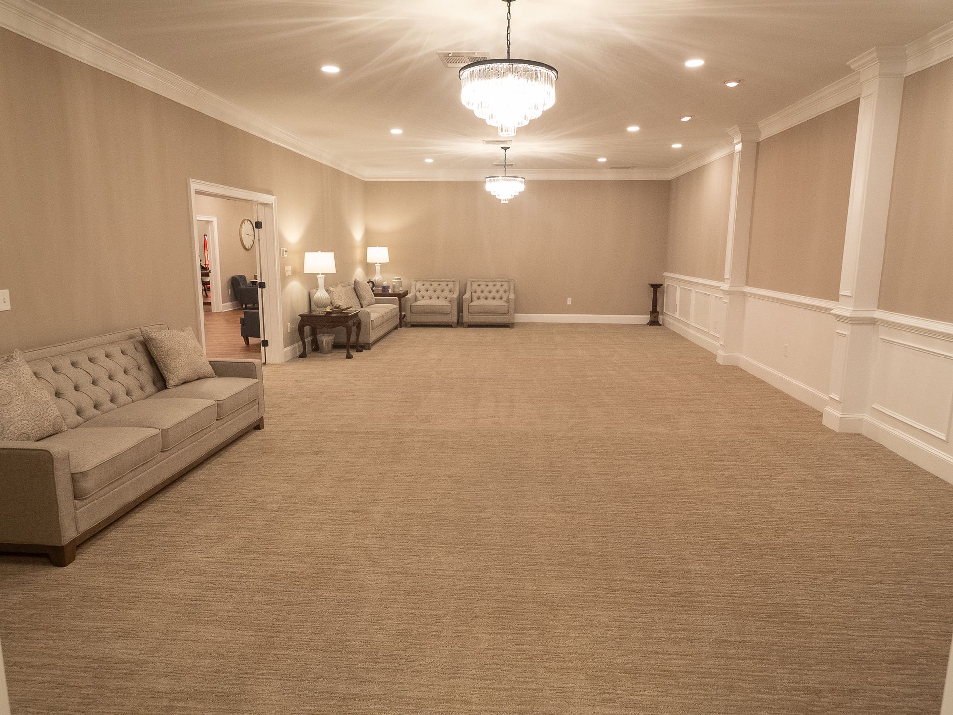 Large beige banquet hall with carpet, chandeliers, chairs, and sofas along the walls.