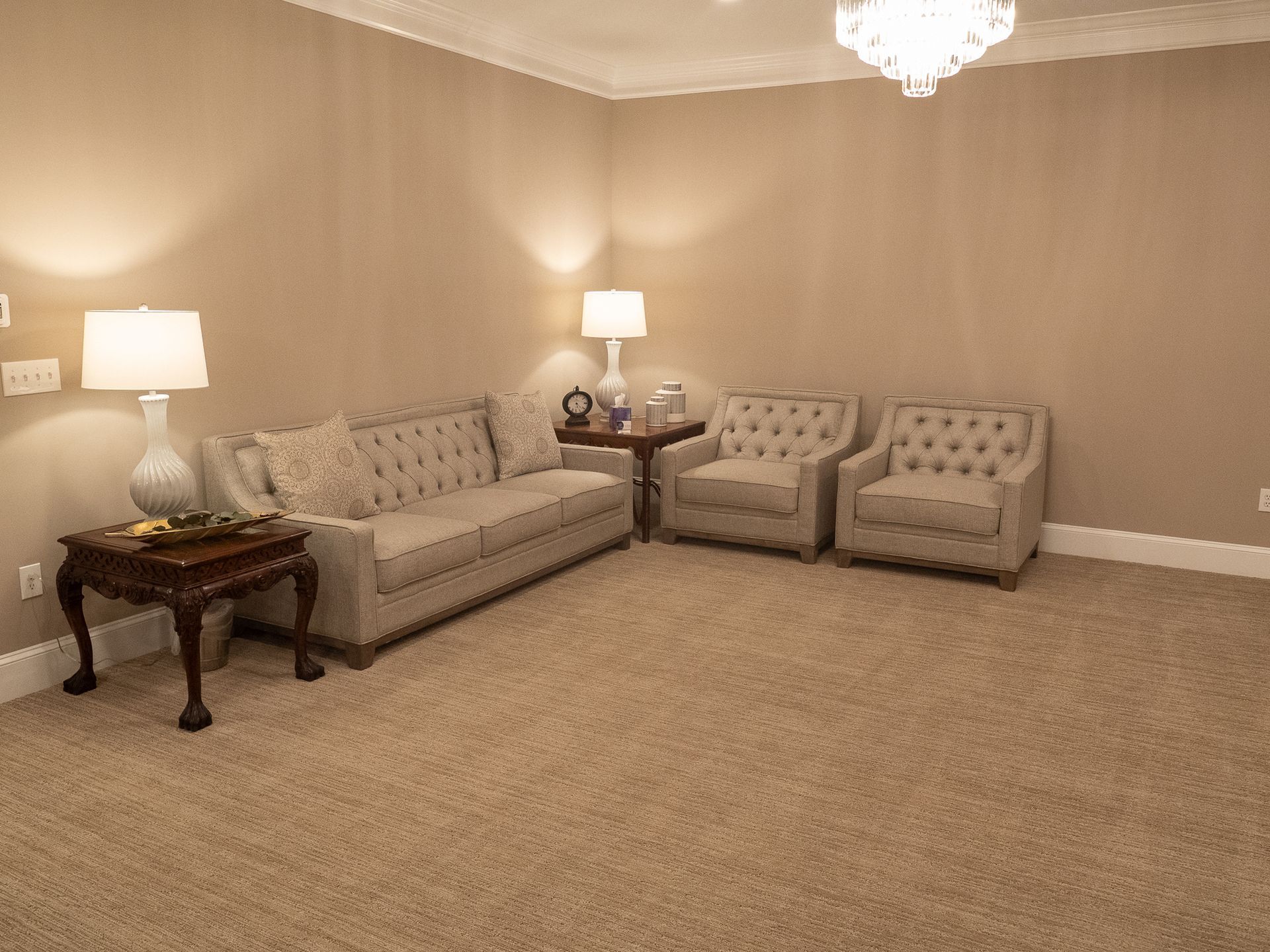 Beige living room with two sofas, two armchairs, side tables, lamps, and carpeted floor.