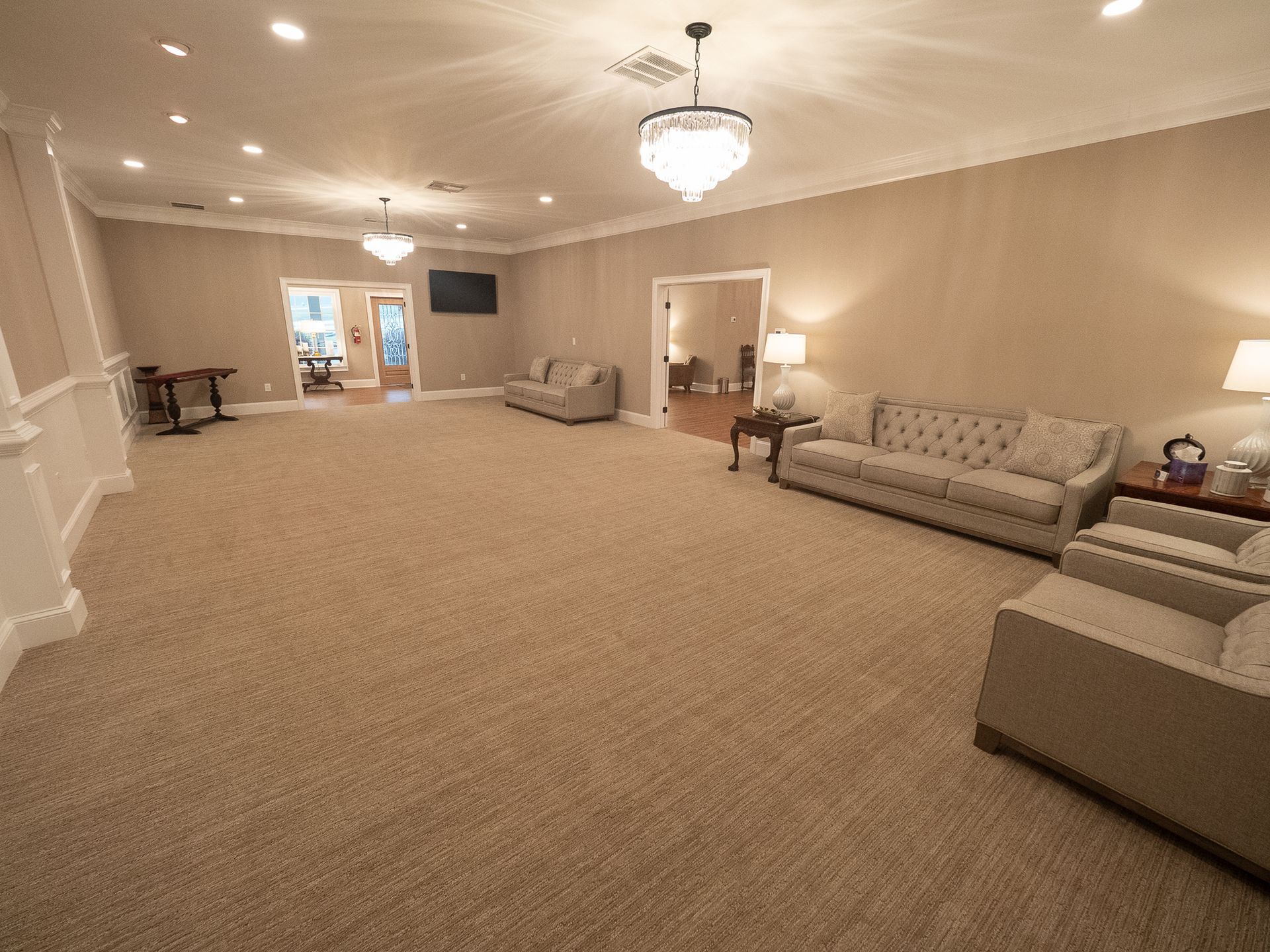 Spacious beige lounge with carpet, sofas, chandelier, and tables near a distant doorway.