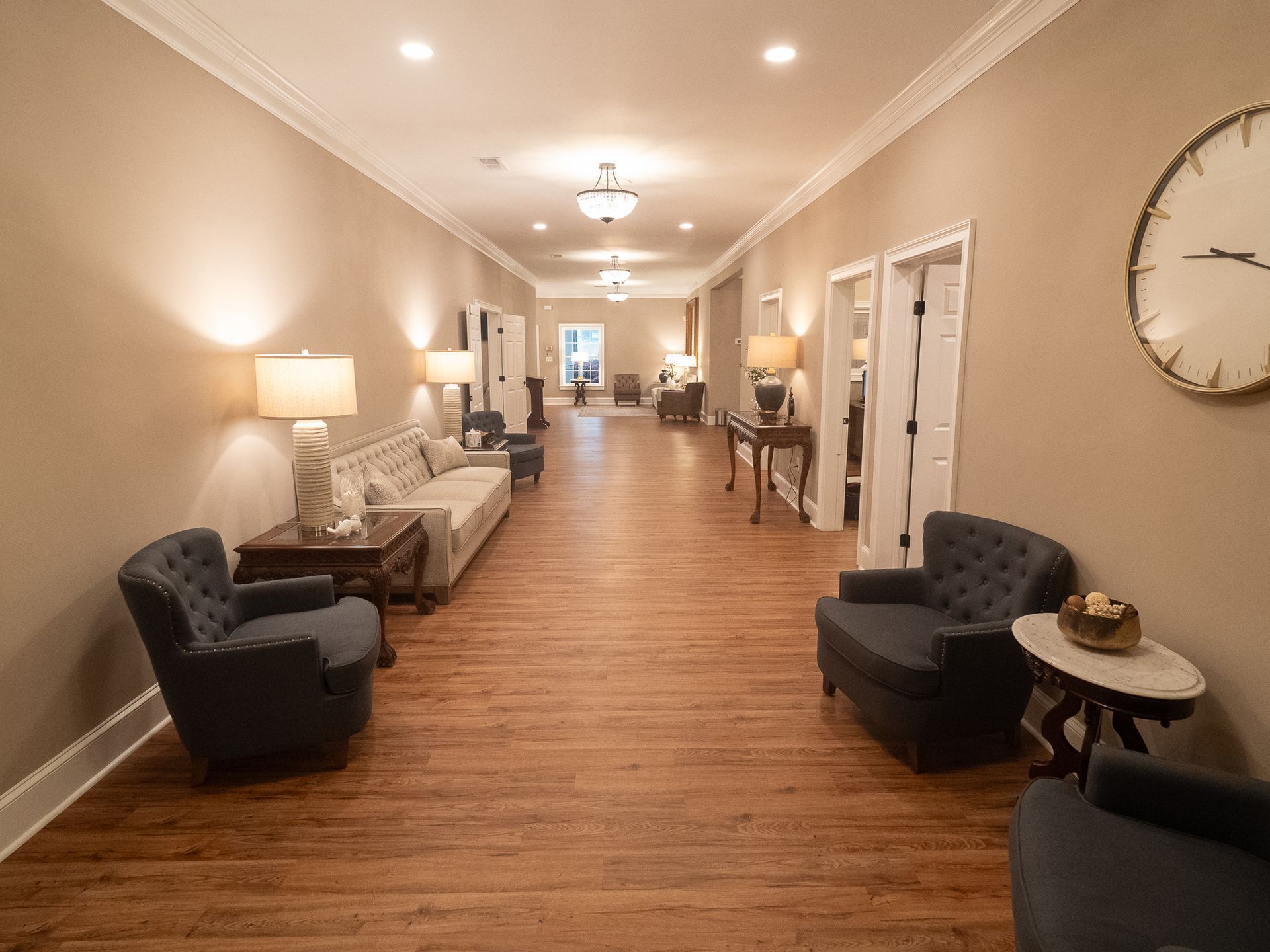 Long beige hallway with wood floors, chairs, lamps, and wall art in a hotel or office lobby