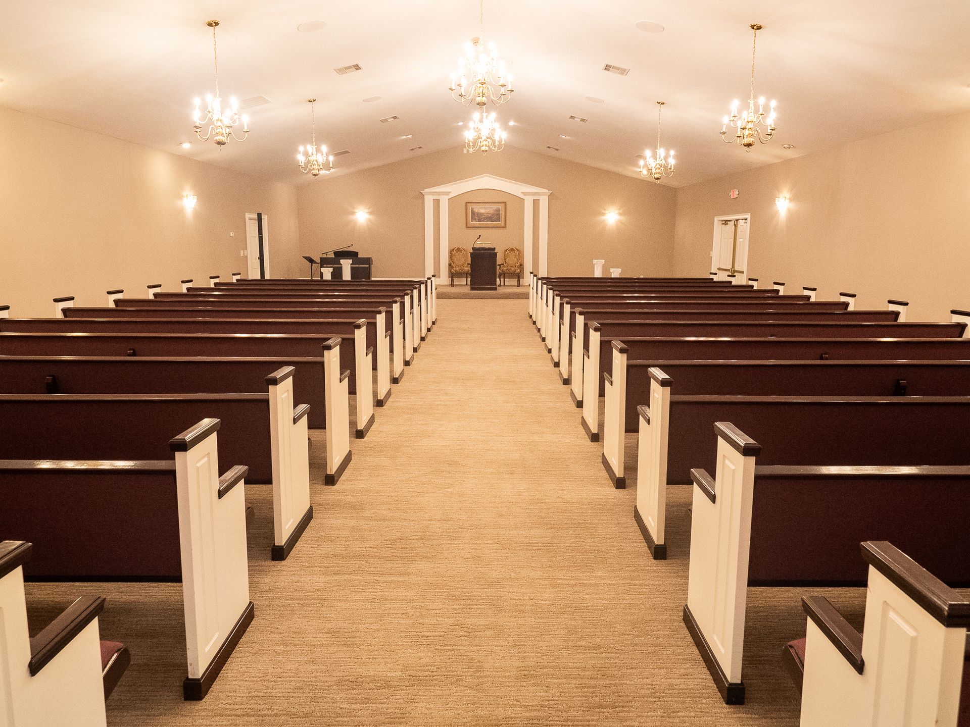 Empty church sanctuary with rows of wooden pews, beige carpet, and chandeliers under a vaulted ceiling