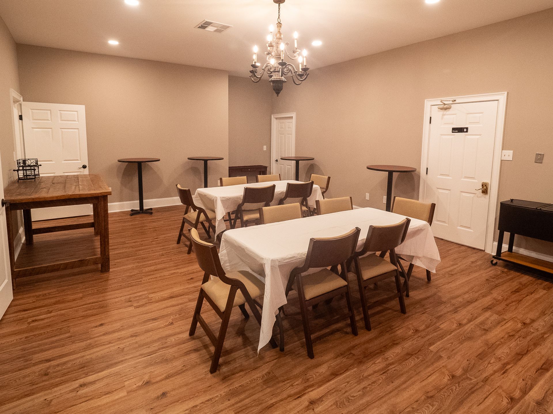 Empty banquet room with round tables, white chair covers, chandeliers, and hardwood floors