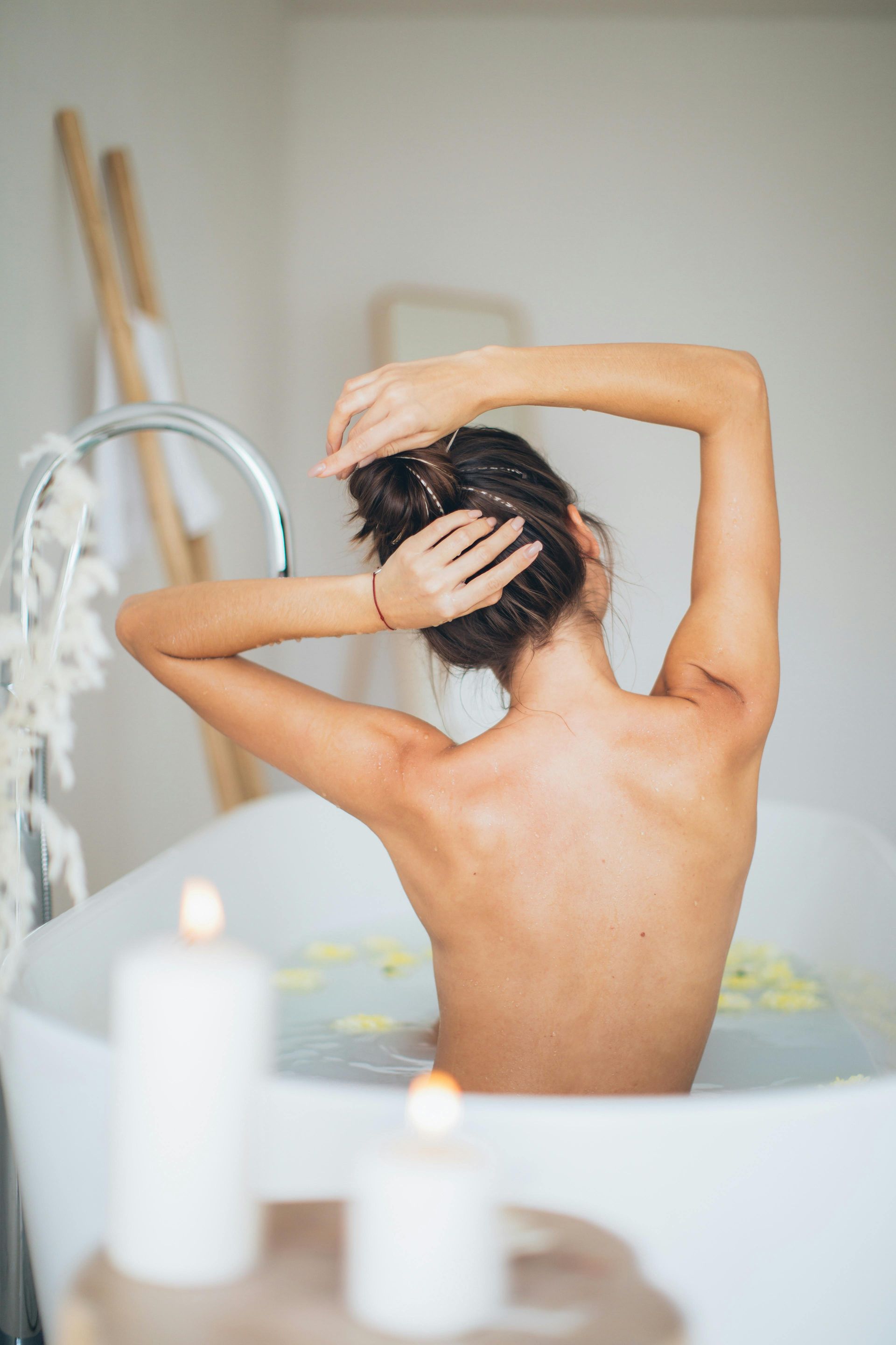 Woman in a white bathtub, reaching up with hands through her hair. Candles and flowers surround.
