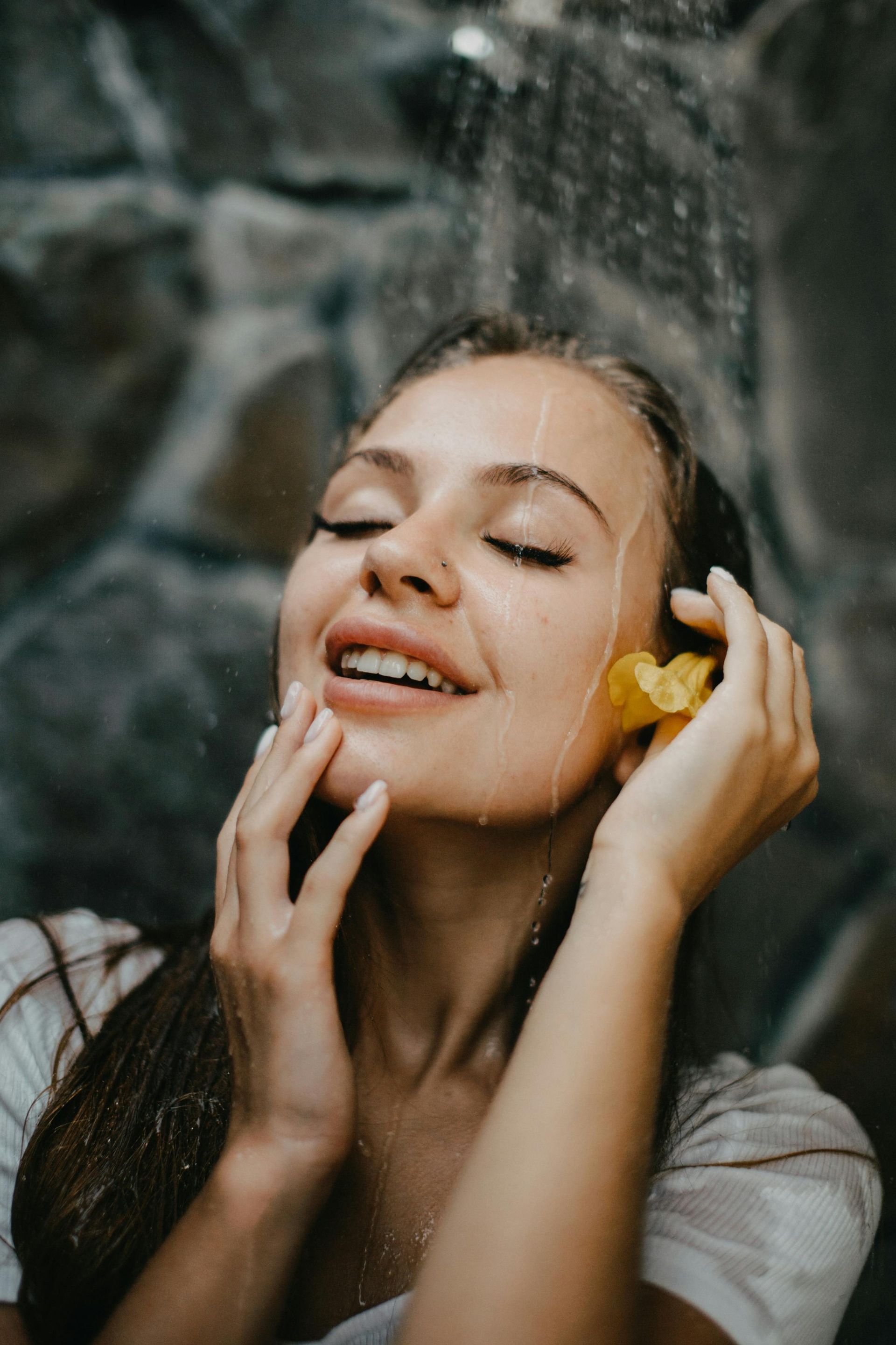 Woman in a shower with eyes closed, smiling, touching her face. Stone wall background, yellow flower.