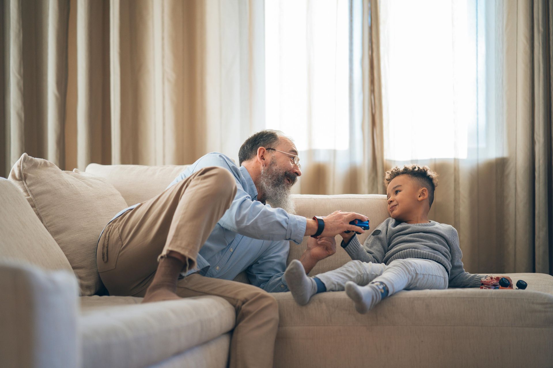 Grandfather and grandson playing on a sofa indoors; grandfather smiles, holding toy.
