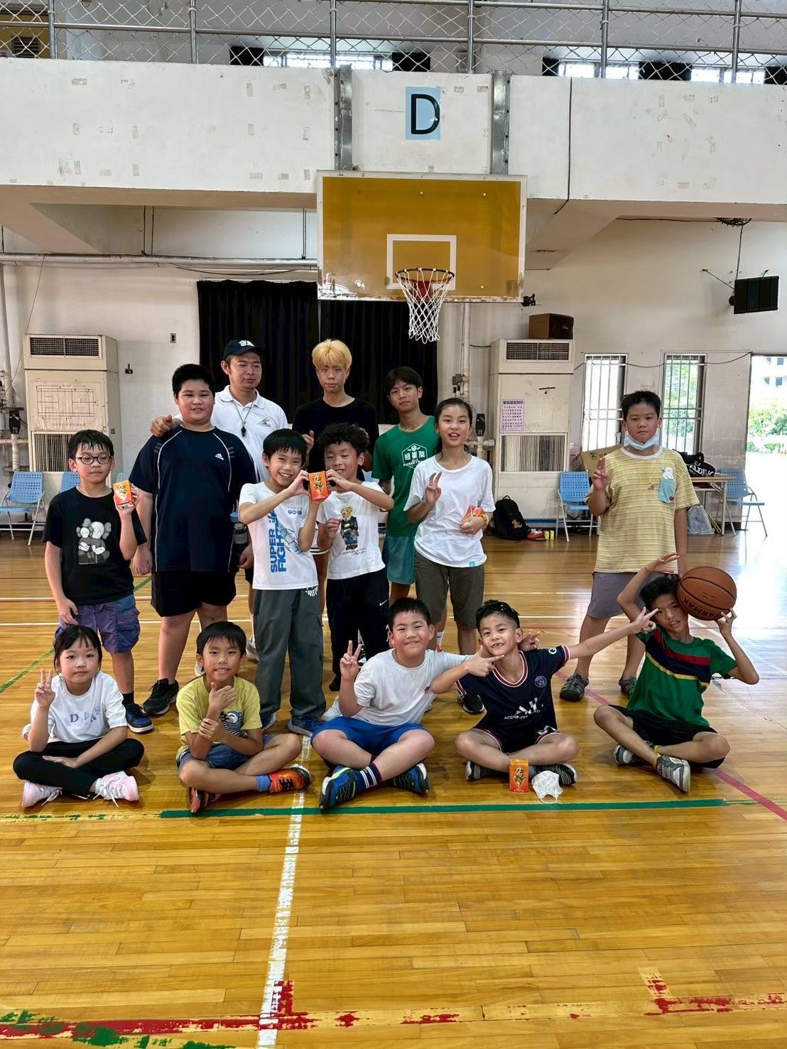 一群小孩和大人在籃球場上擺姿勢拍照。有些人拿著籃球;有些人擺出和平手勢。