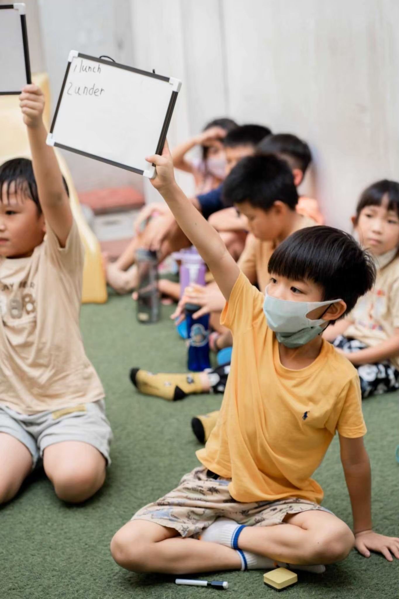 安親班教室裡,孩子們舉著白板,有的戴著口罩,站在綠色的地毯上。