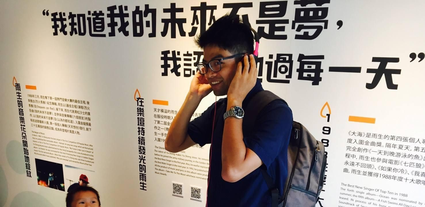 好時光安親班Jack主任戴著眼鏡、戴著粉紅色耳機的男子,在一面寫有中文的牆附近微笑。