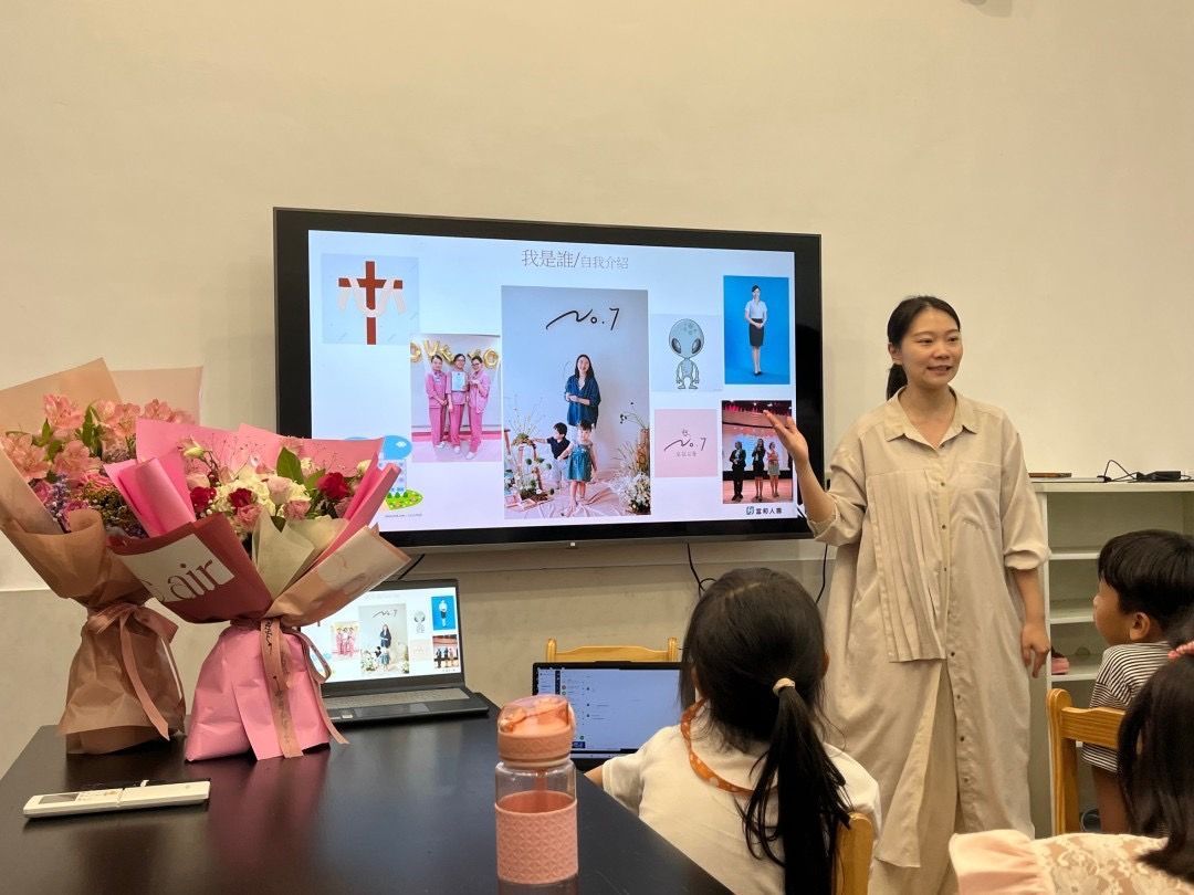 一位女士在教室裡做演講，教室螢幕播放圖片。前景是鮮花和兒童。