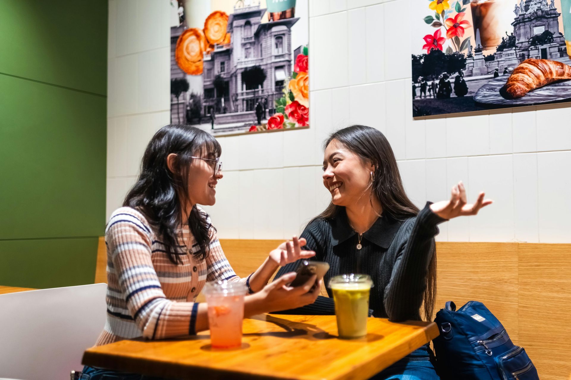兩名女子麵帶微笑，坐在咖啡館裡的一張桌子旁，桌上放著飲料，她們正在看手機。