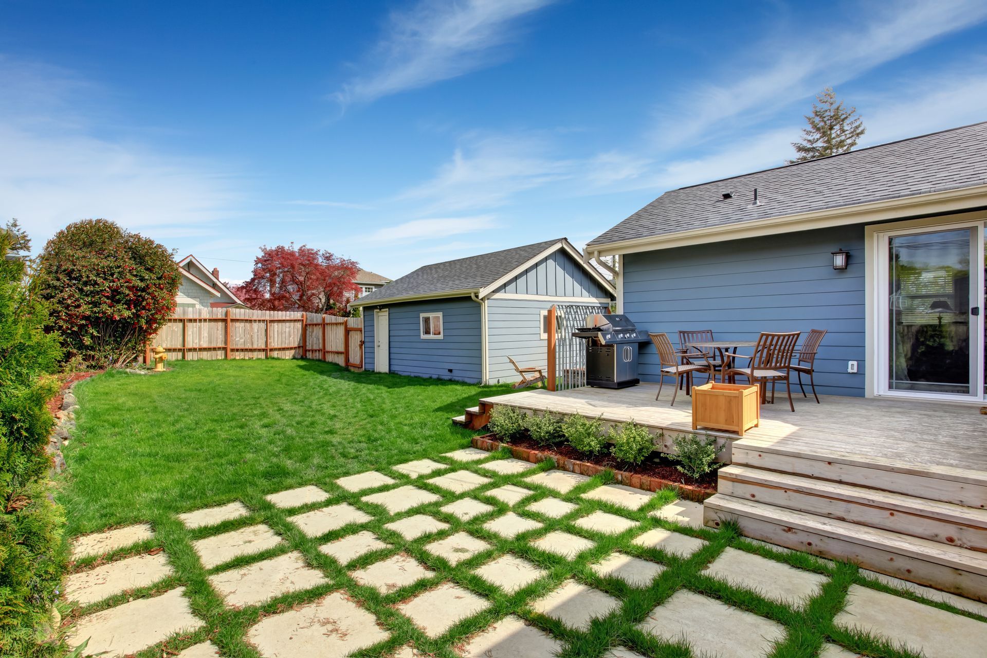A sunny backyard with a blue house, a matching shed, a patio with chairs, and a grid-patterned stone path on green grass.