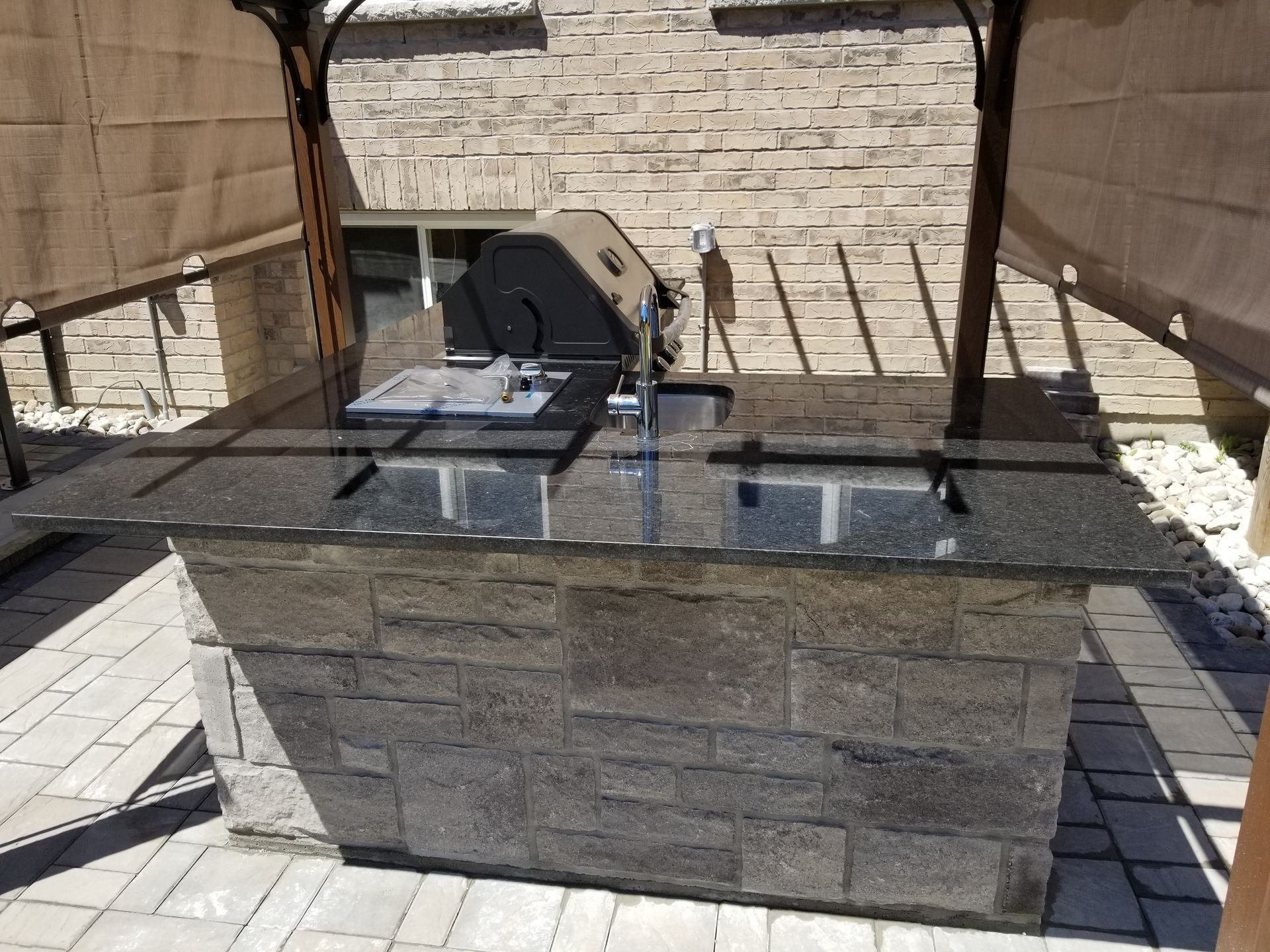 Outdoor stone-faced kitchen island with a grill and sink, topped with black granite. Beneath a shaded pergola.