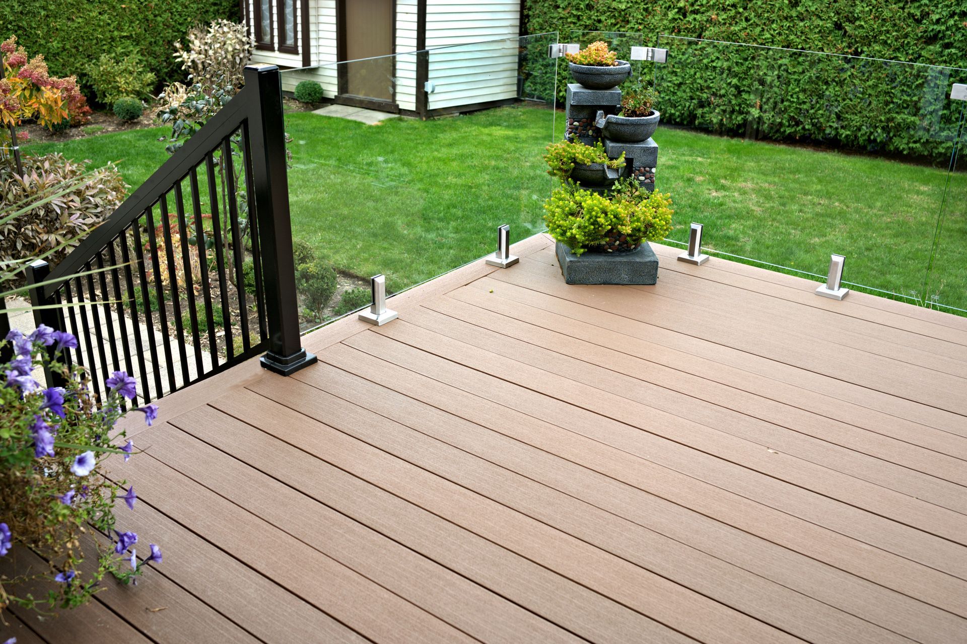 A wooden deck with a glass railing, black metal stair handrail, and a tiered fountain, overlooking a green lawn.