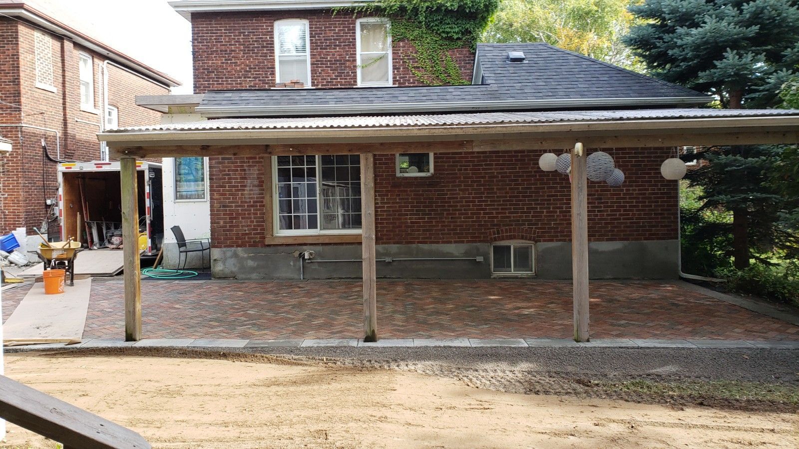 A brick house with a paved patio covered by a wooden pergola, viewed from a sandy yard.