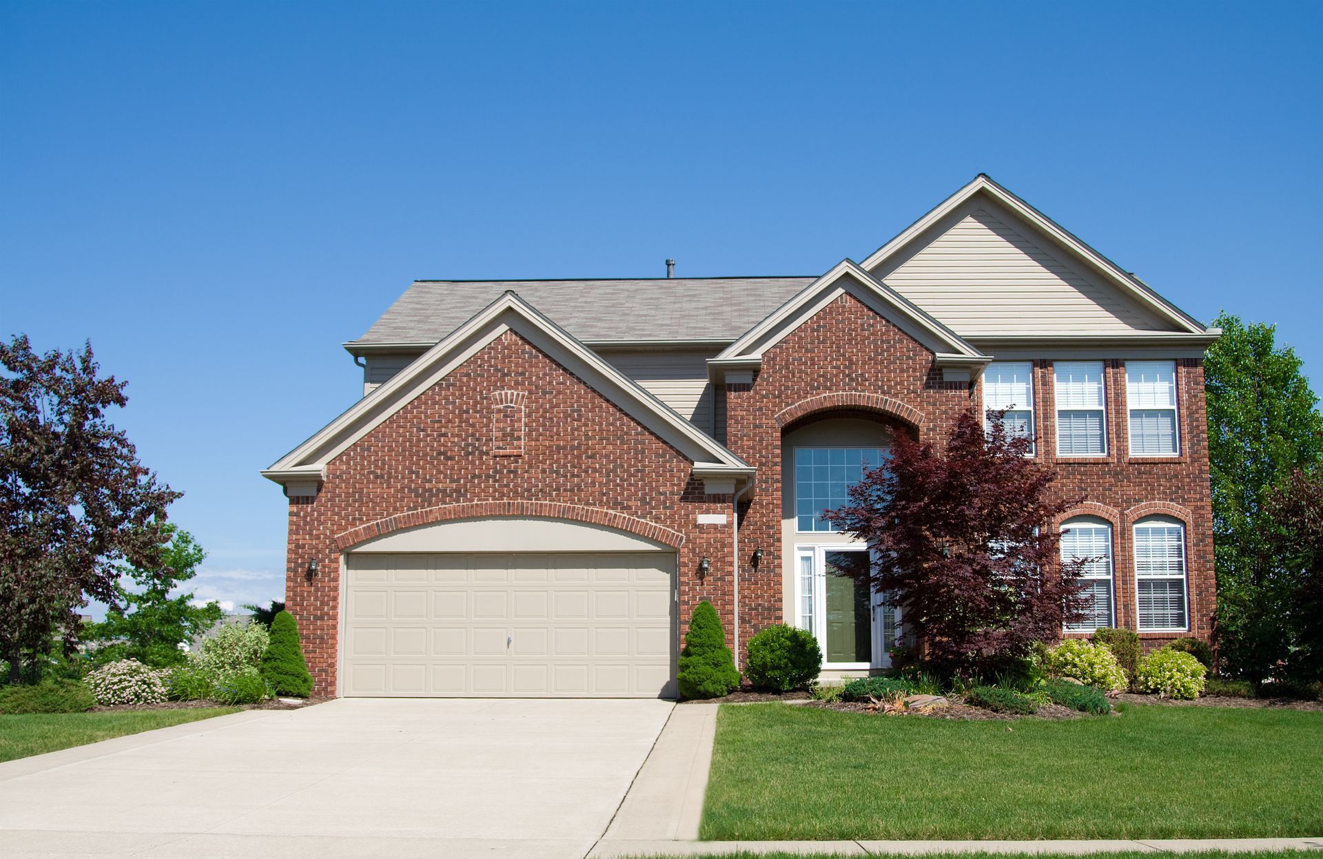 Brick suburban house with a two-car garage, driveway, and green lawn under a clear blue sky
