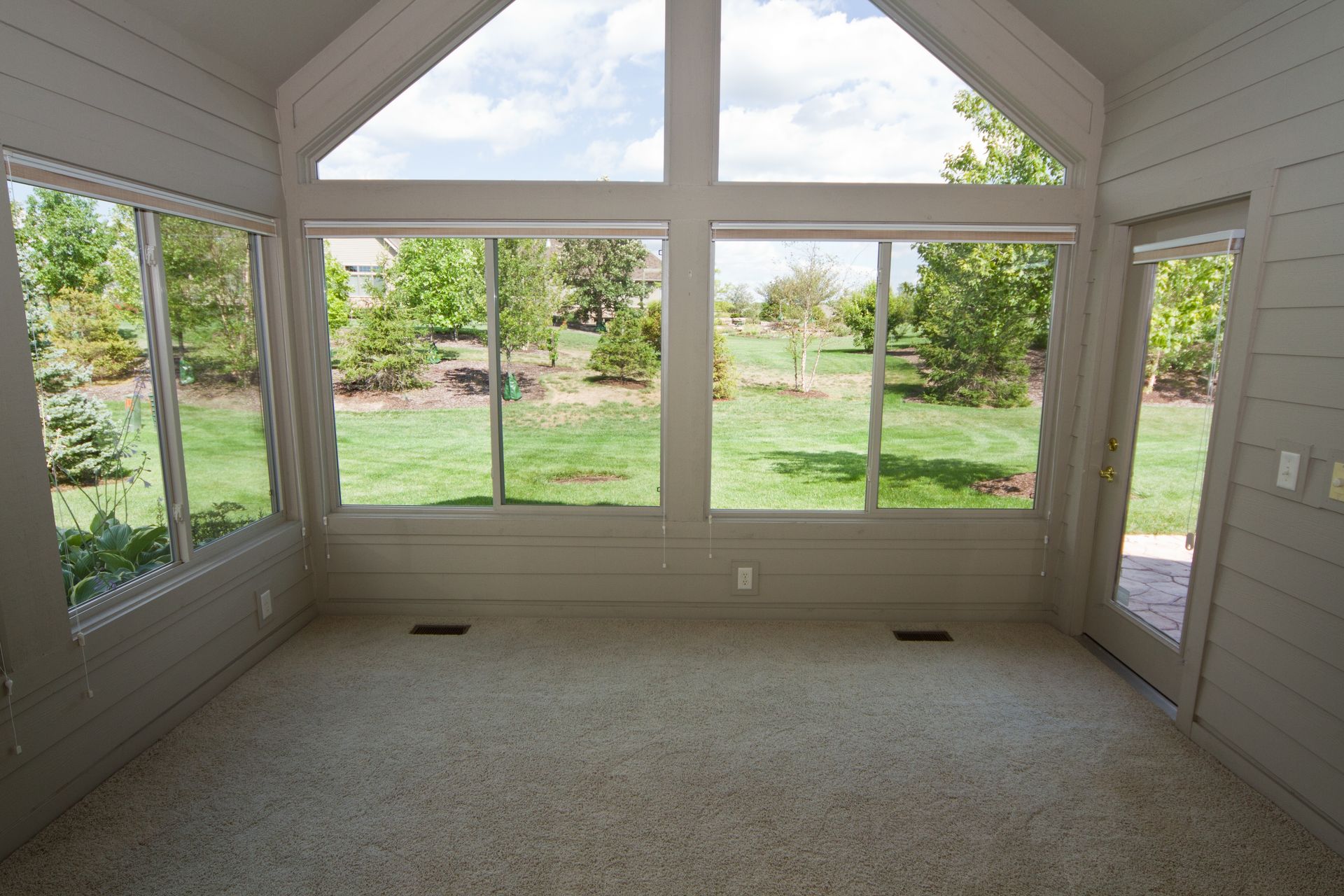 A sunroom with beige walls, gray carpeting, and large windows overlooking a grassy backyard with trees.