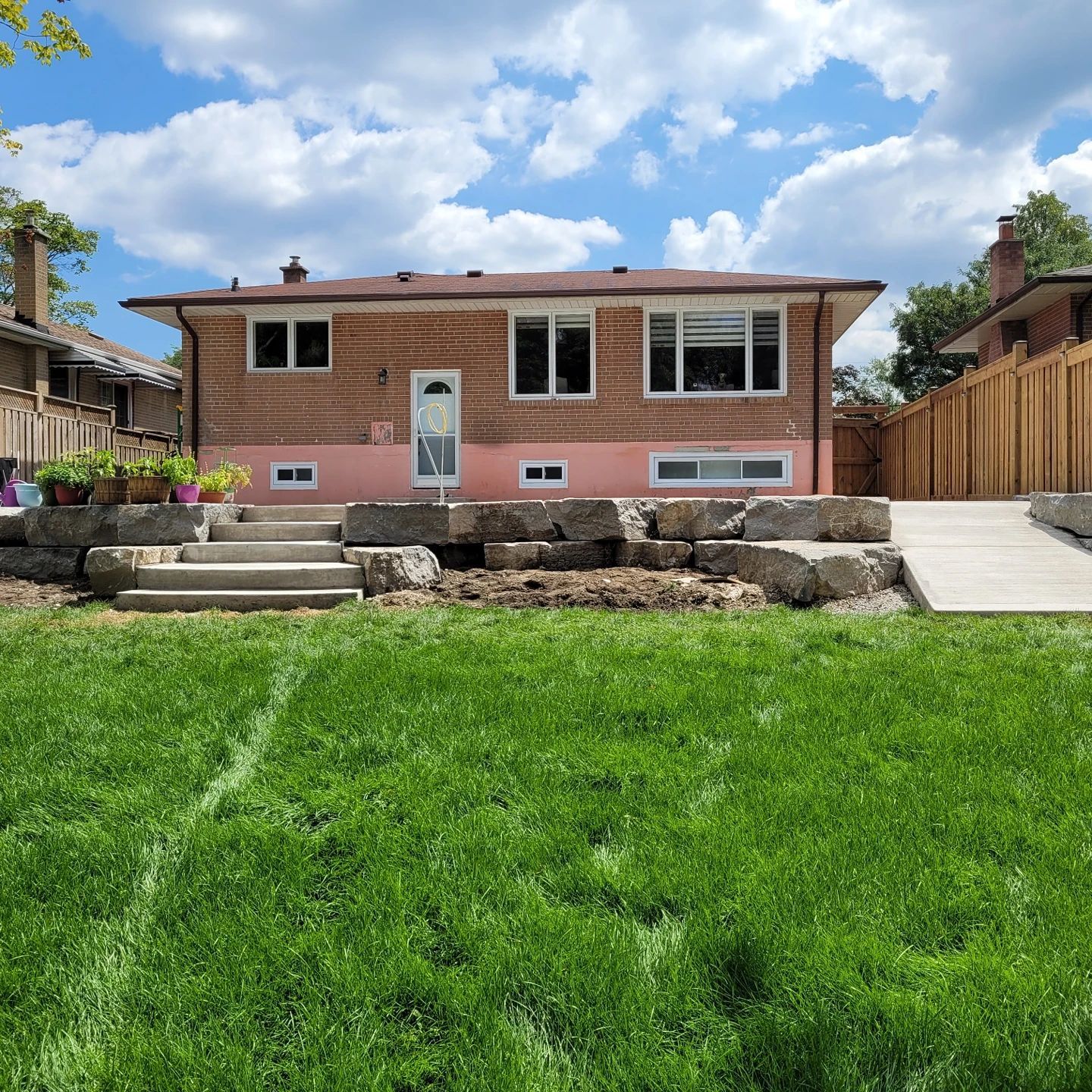 Back view of a brick house with a pink foundation, stone retaining wall, concrete steps, and a lush green lawn.