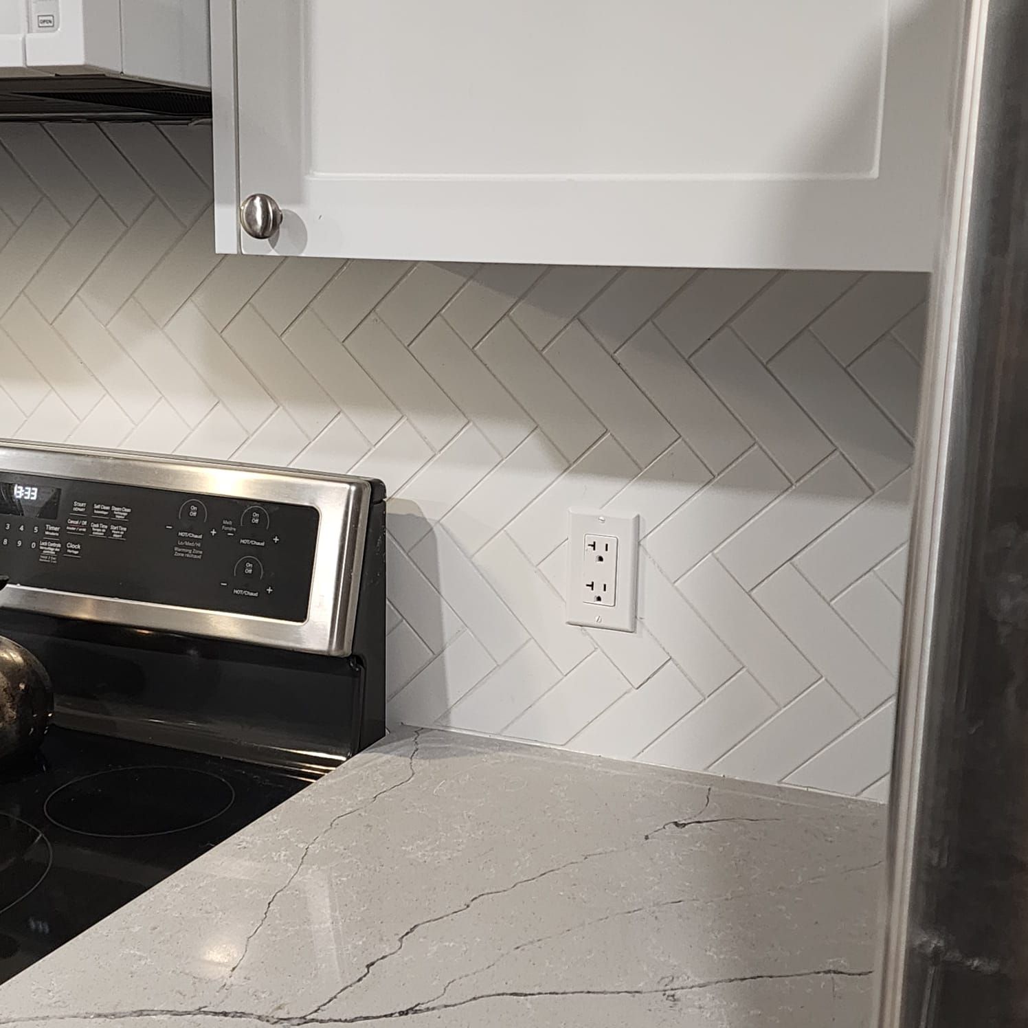 White kitchen backsplash with herringbone pattern, electrical outlet, and white cabinets.