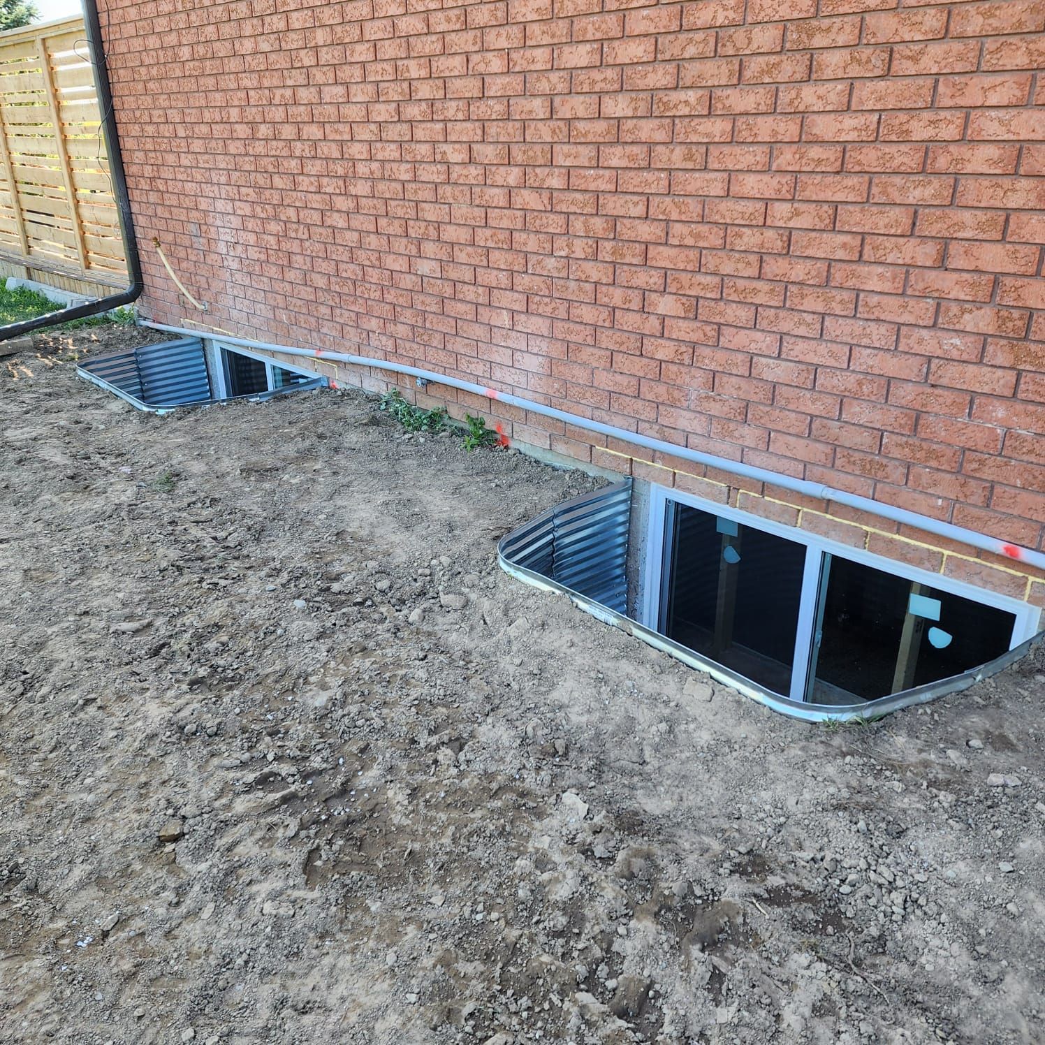 Brick wall with two basement window wells in dirt.