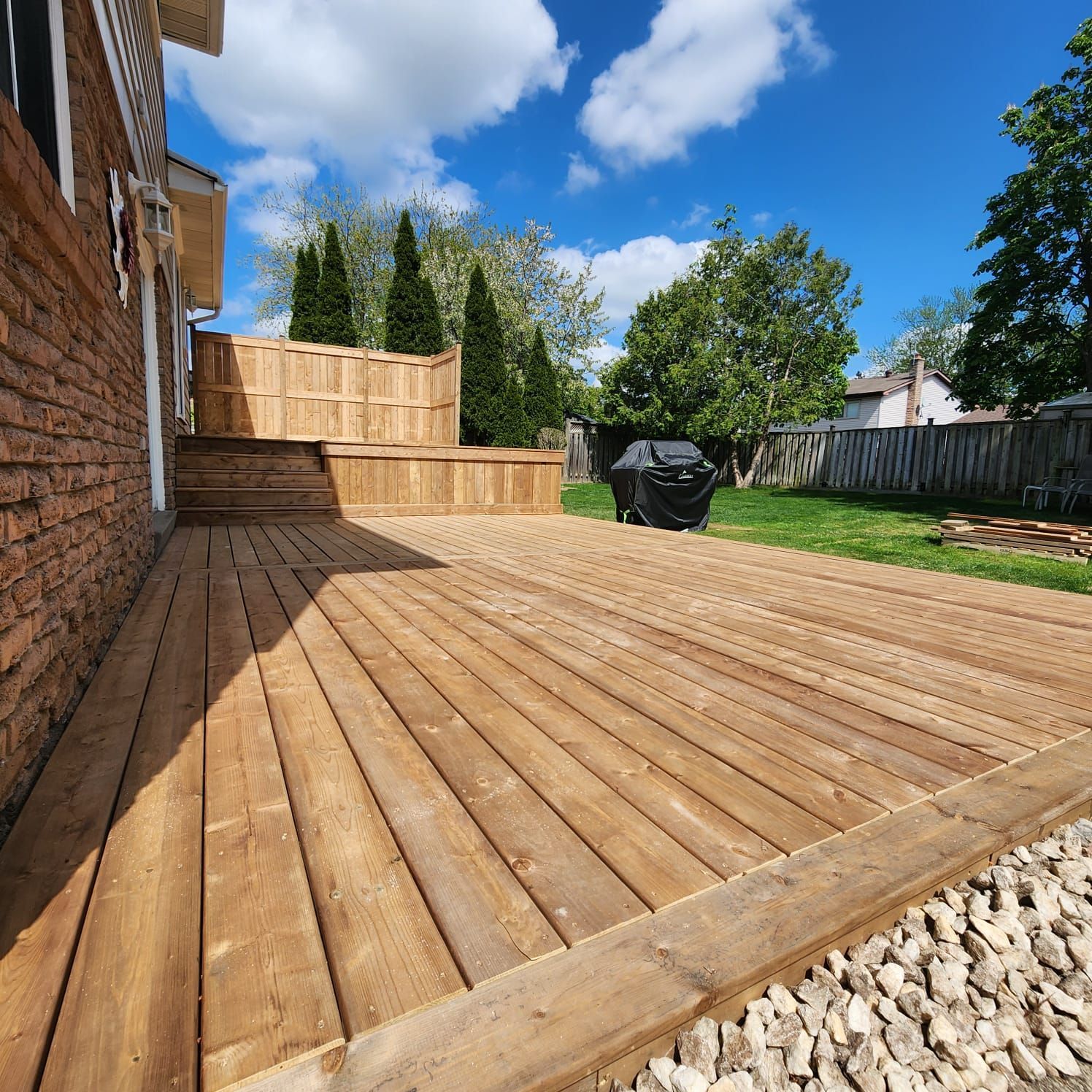 Wooden deck with raised platform, brick house side, and a sunny backyard with trees and a grill.
