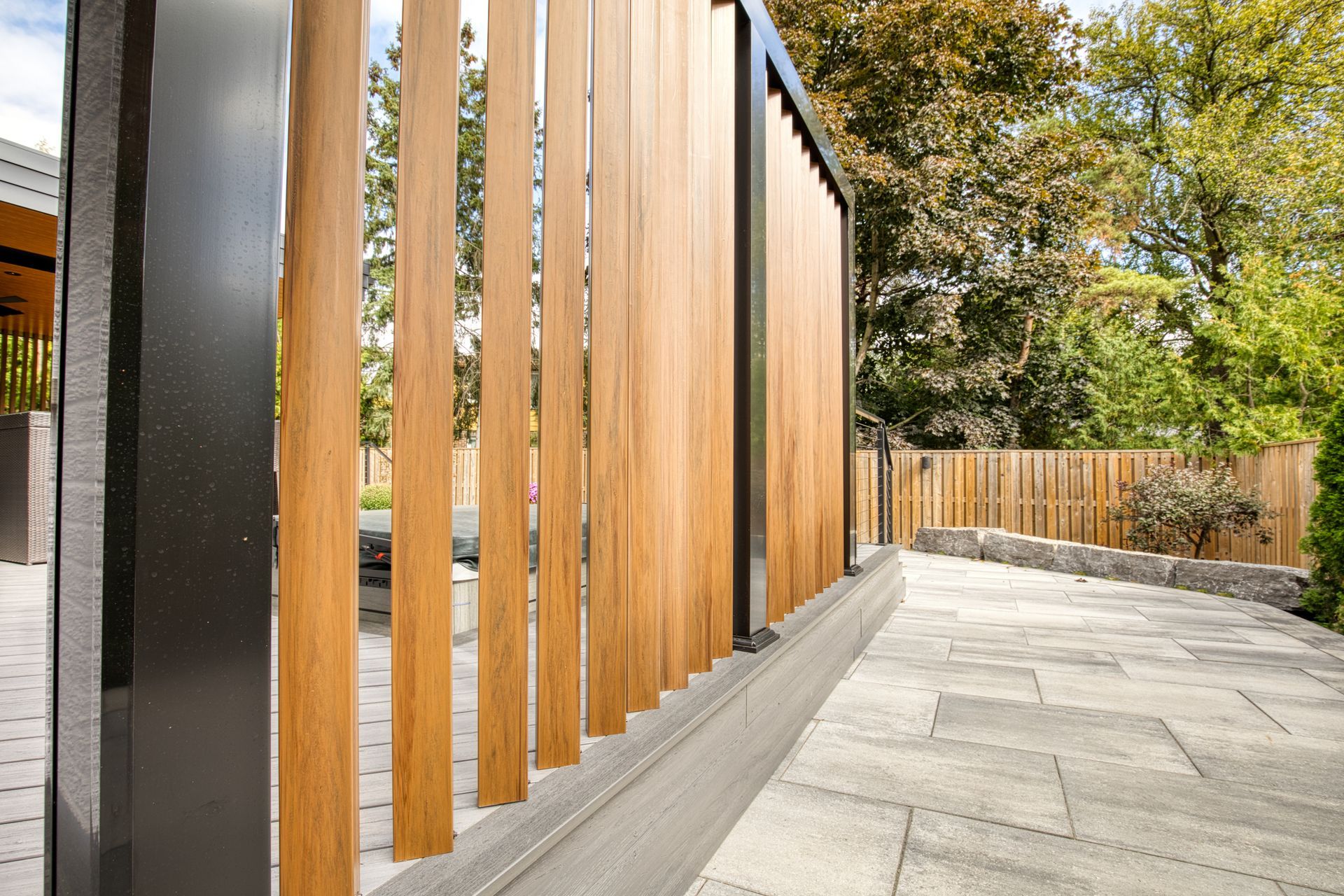 Wooden slat fence with black frame, next to stone walkway, with trees in the background.