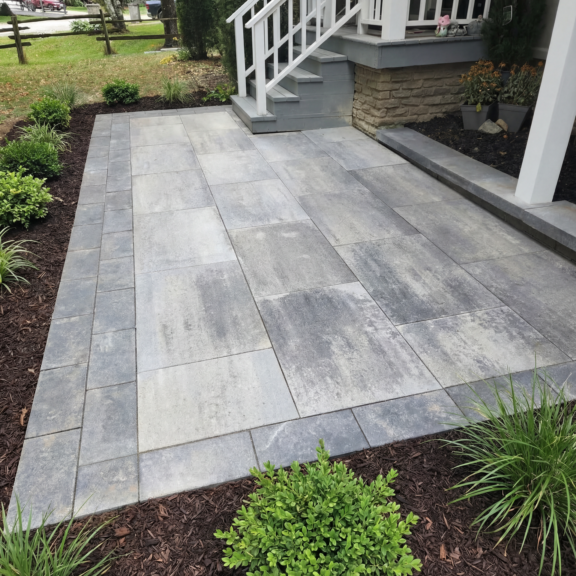 Paver patio with a dark border in front of a house entrance. Greenery surrounds it on mulch.