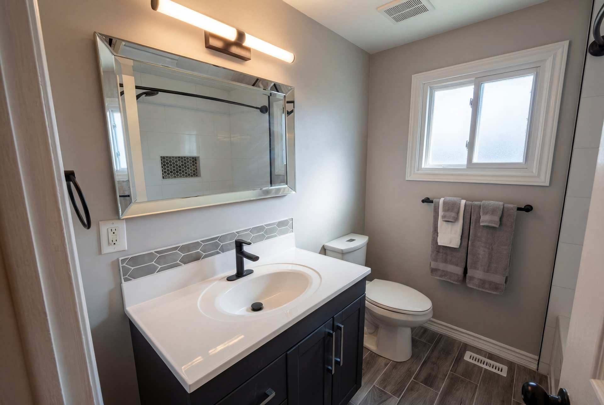 Bathroom with vanity, mirror, toilet, and window; gray walls, white countertop, and dark cabinet.